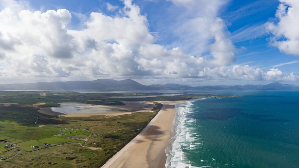A coastal landscape with a beach, farmland, and distant mountains under a partly cloudy sky.