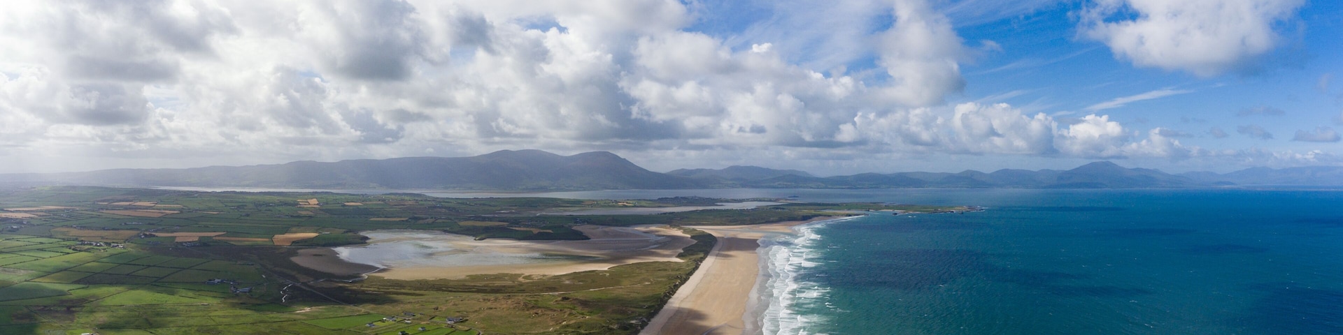 A coastal landscape with a beach, farmland, and distant mountains under a partly cloudy sky.