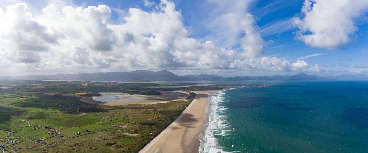 A coastal landscape with a beach, farmland, and distant mountains under a partly cloudy sky.
