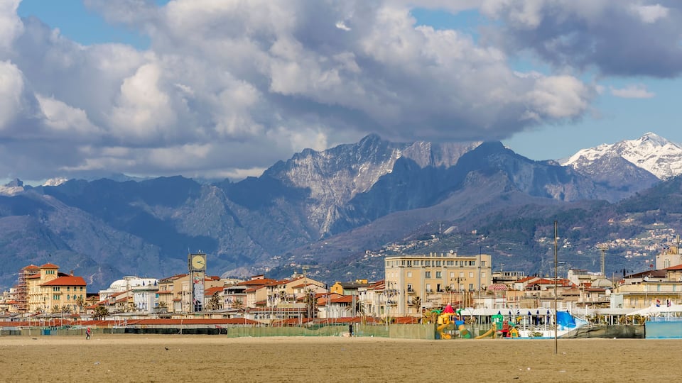 A coastal town with a prominent clock tower, colorful buildings, and snow-capped mountains in the background.
