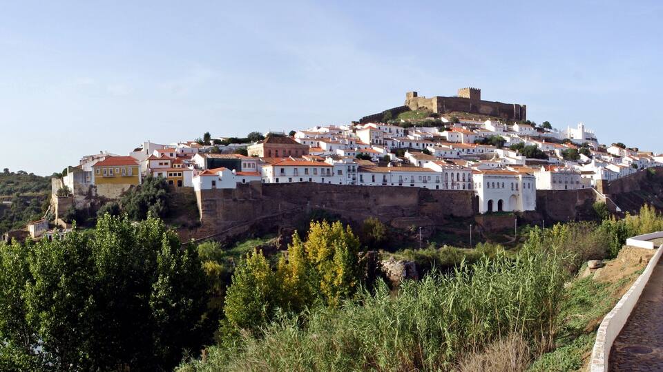 A historic town with white buildings on a hill, a river in the foreground, and a castle on top.