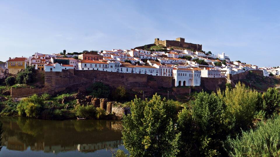 A hilltop town with white buildings, a river in the foreground, and a castle on top.
