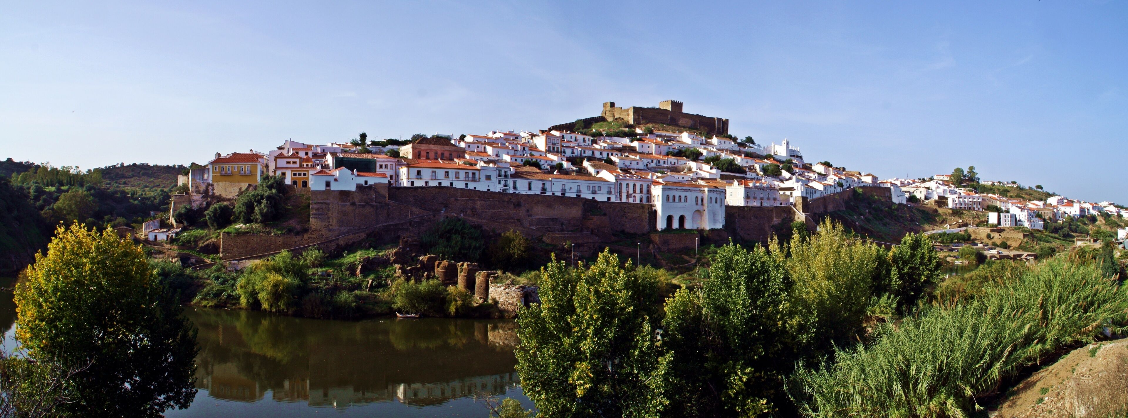 A hilltop town with white buildings, a river in the foreground, and a castle on top.