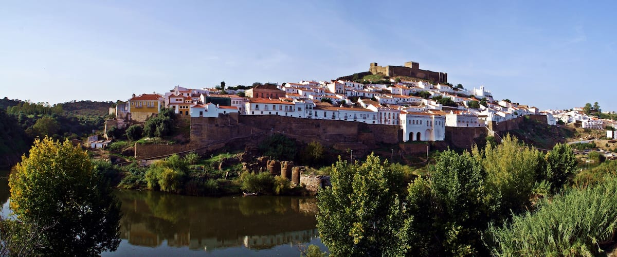 A hilltop town with white buildings, a river in the foreground, and a castle on top.