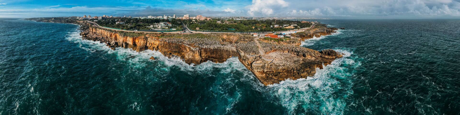 A coastal cityscape with a prominent rocky peninsula, a bridge connecting to the mainland, and a clear blue sky.