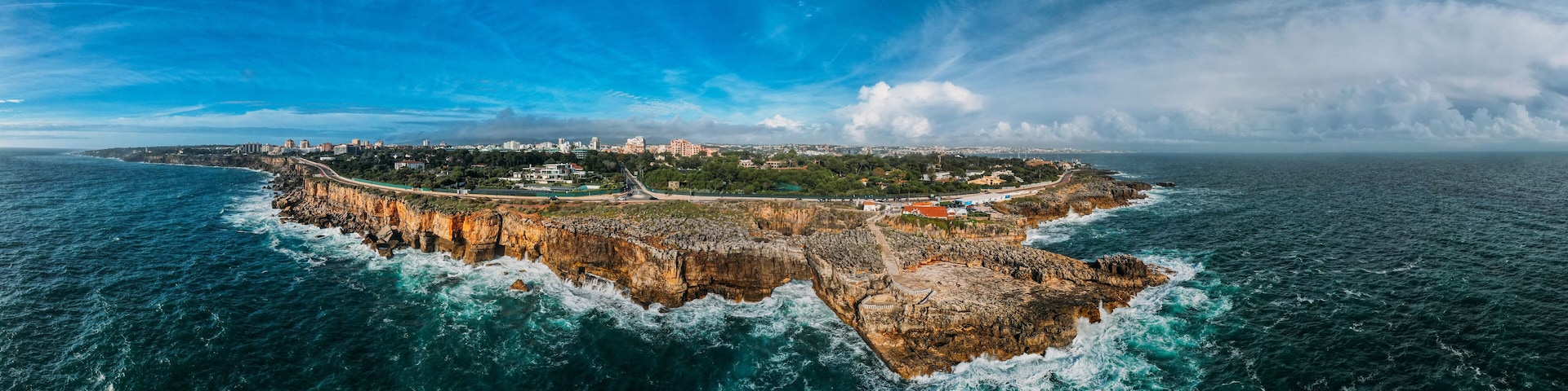 Un paysage urbain côtier avec une péninsule rocheuse imposante, un pont reliant la ville au continent, et un ciel bleu dégagé.