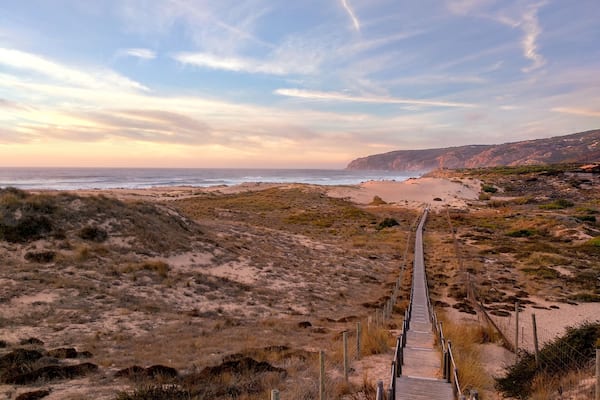 Eine Küstenlandschaft mit einem hölzernen Steg, der zu einem Strand führt, umgeben von Dünen und Klippen.
