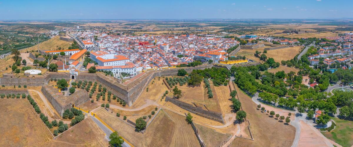 An aerial view of a historic town with a prominent fortification, surrounded by rolling hills and fields.
