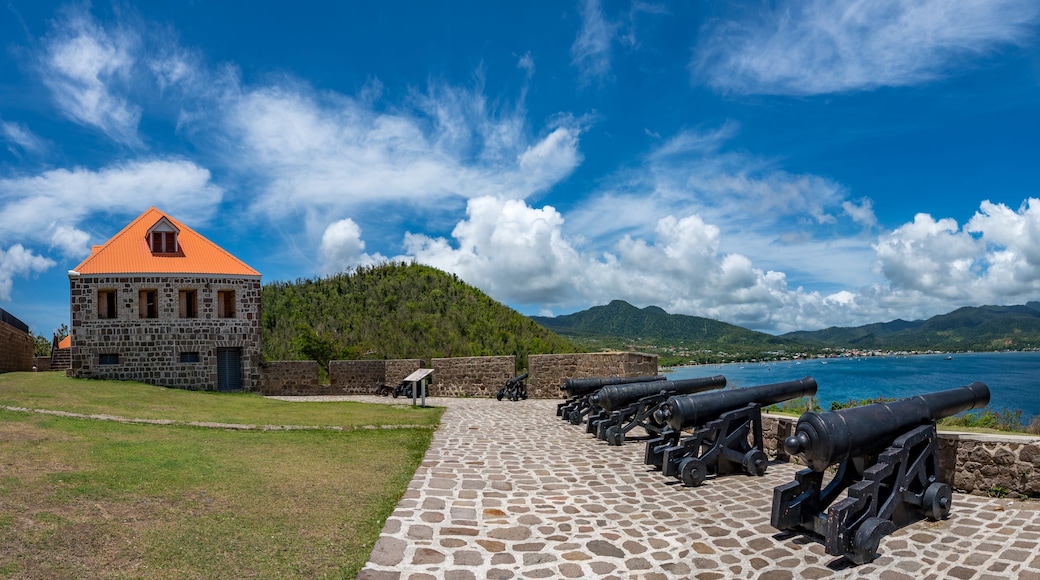 A stone building with a red roof, a row of cannons, and a coastal landscape with mountains in the background.