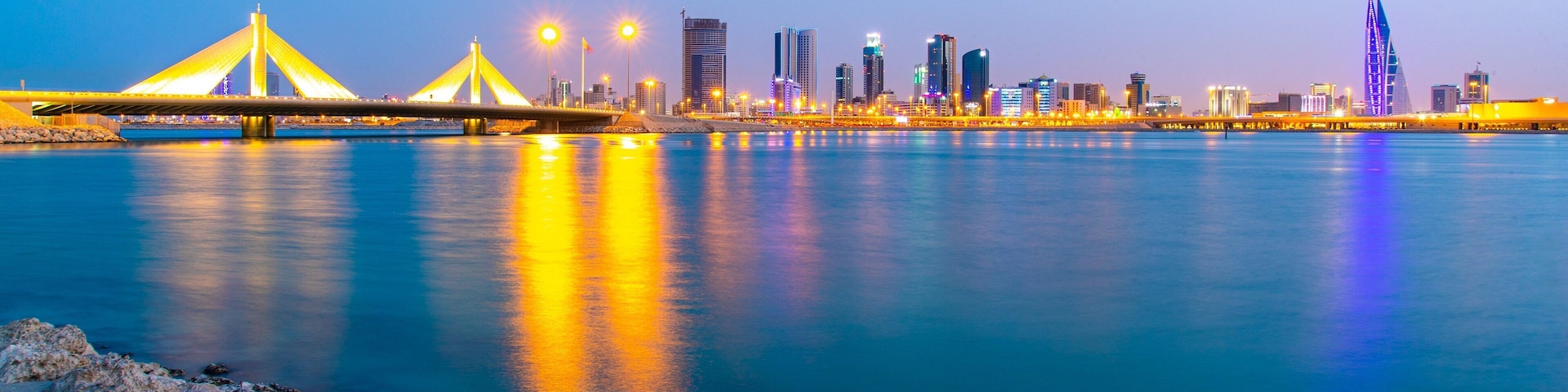 Un horizonte urbano con edificios iluminados y un puente sobre un río tranquilo al atardecer.
