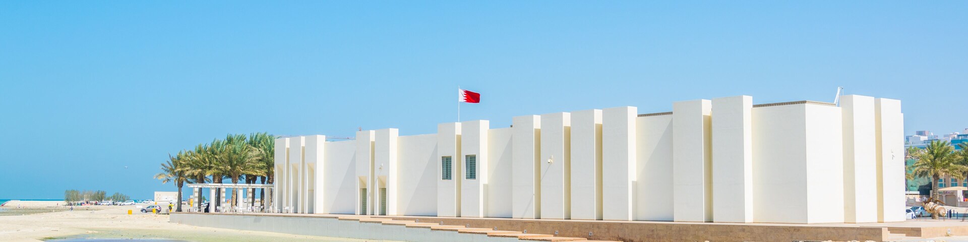 A beach with clear waters, a white building with vertical lines, and a flag.