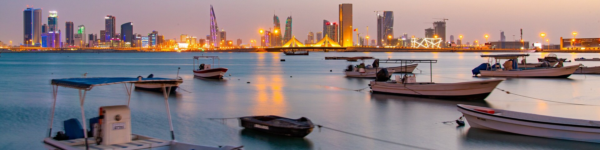 Um cais tranquilo ao entardecer com vários barcos ancorados perto da margem, silhueta urbana ao fundo e uma ponte ligando duas partes da cidade.