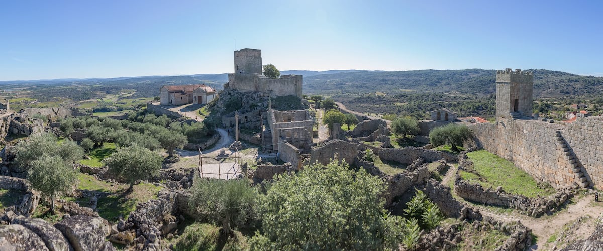 A historical site with ruins, including a tower and walls, surrounded by olive trees and a hilly landscape.