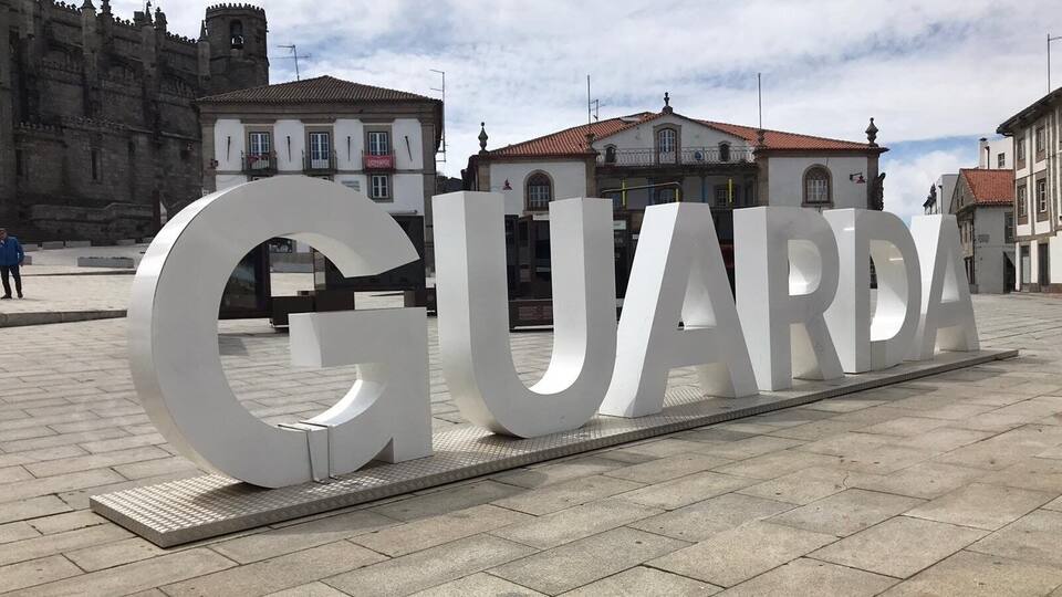 A large white 'GUARDA' sign in a public square with historic buildings in the background.