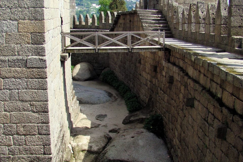 A stone bridge over a moat, part of a historic castle with battlements and a view of the surrounding landscape.