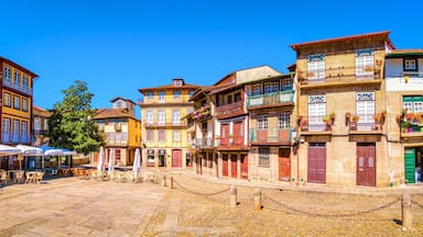 Uma praça de calçada antiga com edifícios coloridos, mesas ao ar livre e um céu azul claro.