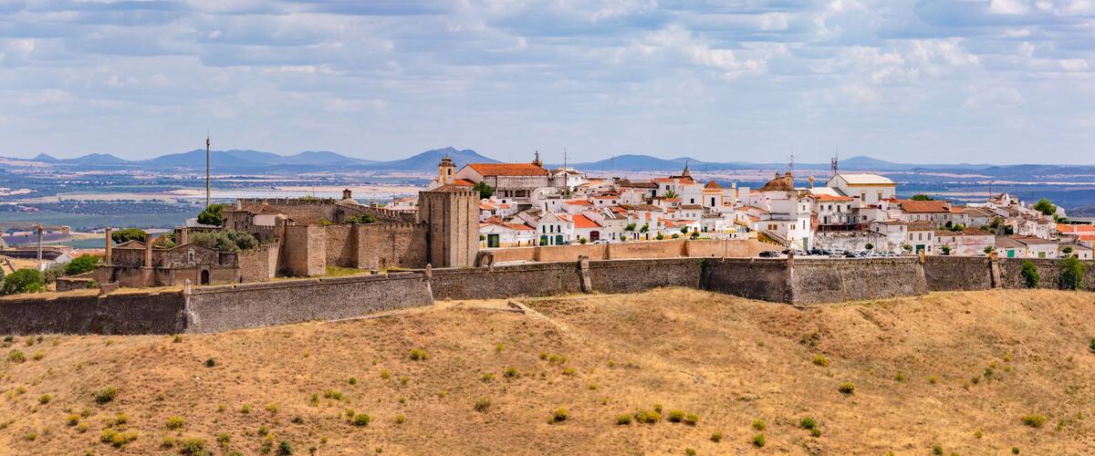 A historic town with a fortified wall, white buildings with red roofs, and a mountainous backdrop.