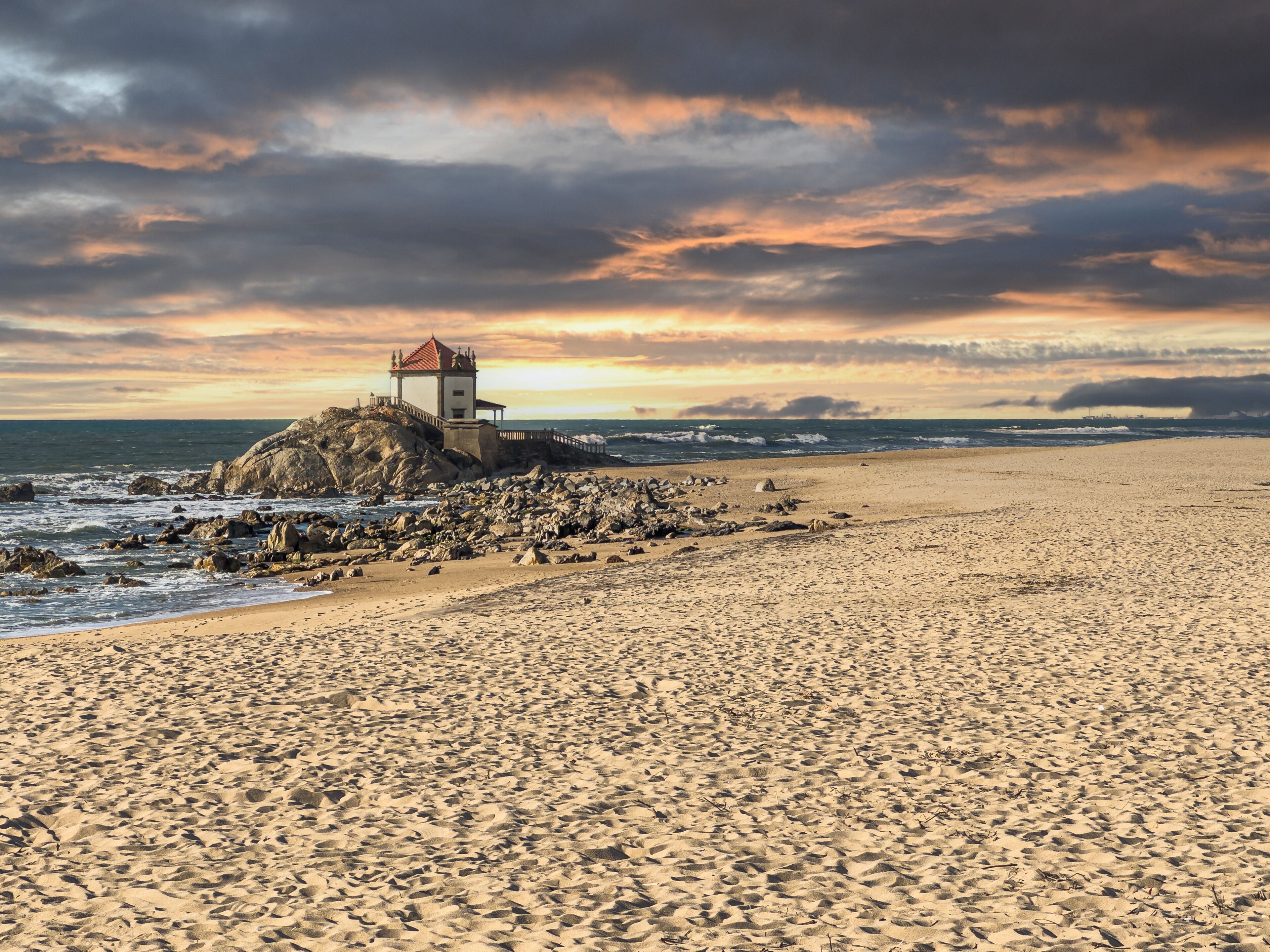 A rocky beach with a lighthouse on a cliff, waves crashing, and a sunset sky.