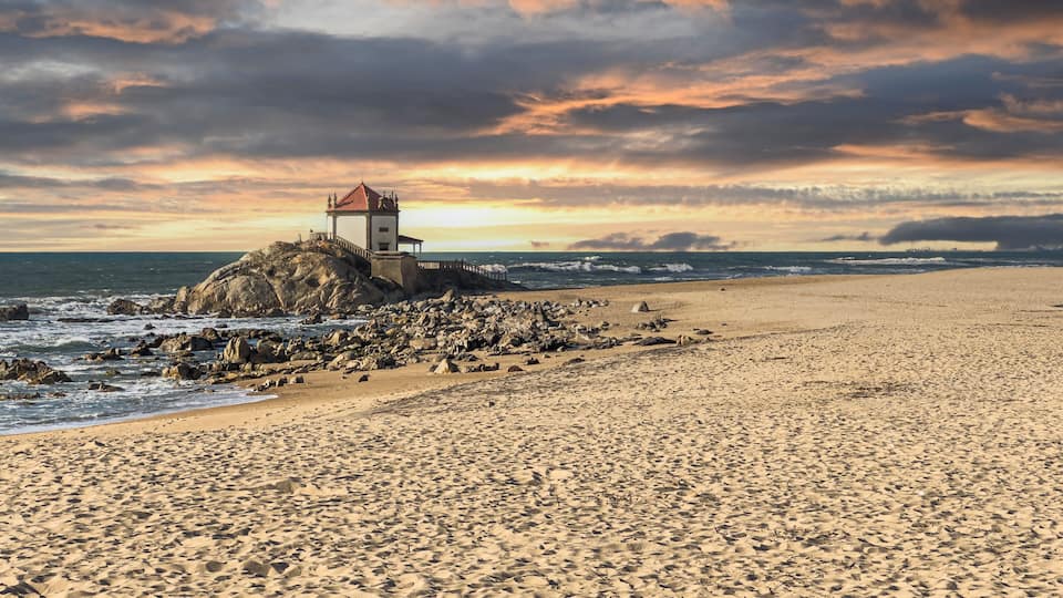 A rocky beach with a lighthouse on a cliff, waves crashing, and a sunset sky.
