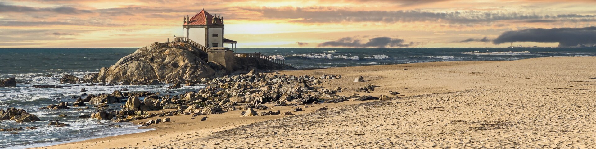 Uma praia rochosa com um farol no alto de um penhasco, ondas quebrando e um céu ao pôr do sol.