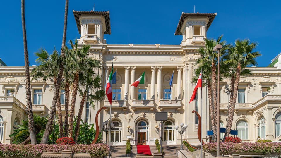 A grand building with columns, palm trees, and multiple flags.