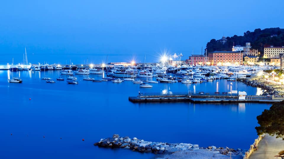 A marina at dusk with numerous boats docked, a rocky shoreline, and buildings along the coast.