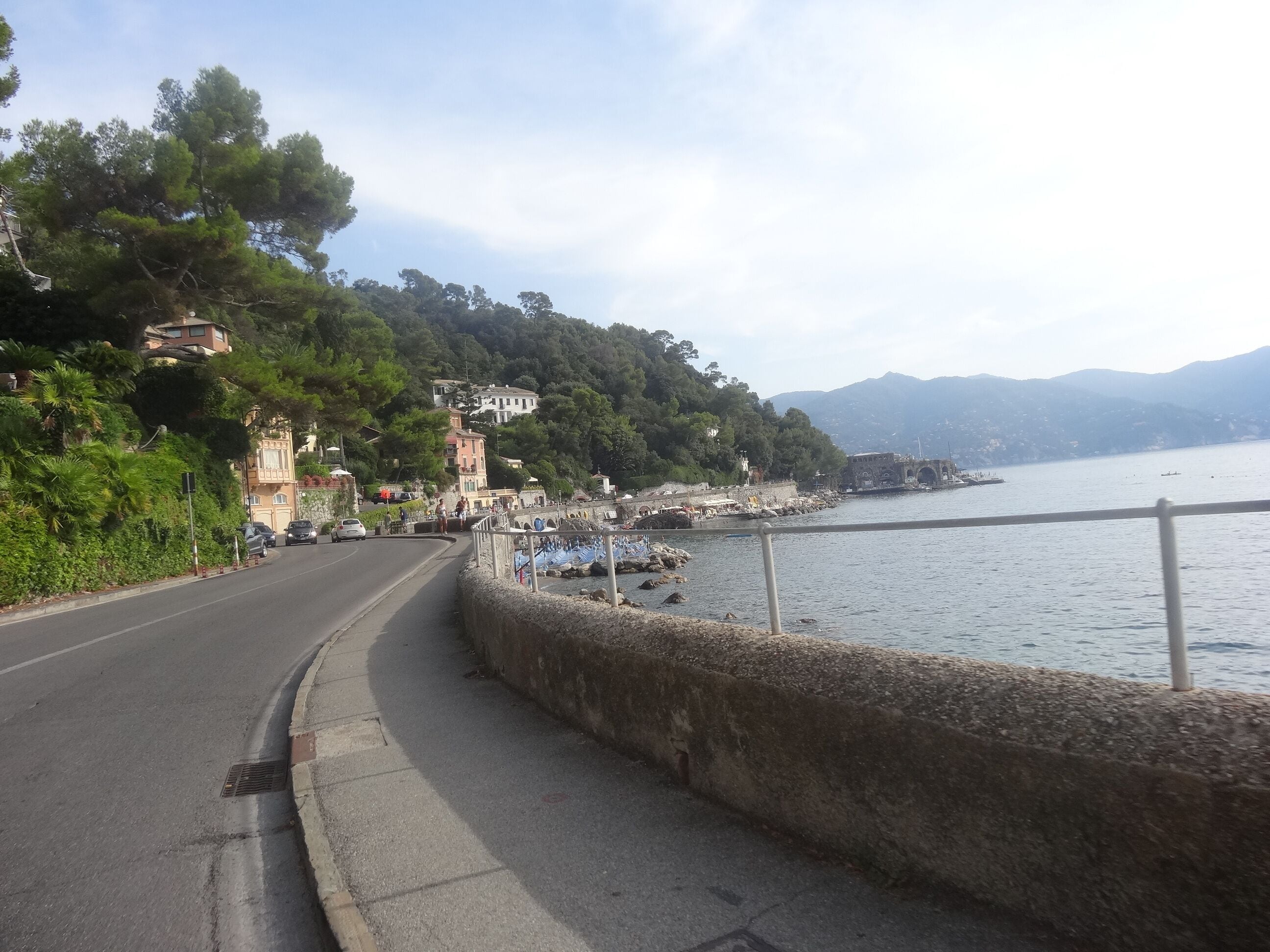 A coastal road with a stone wall, a bridge, and a body of water with boats.