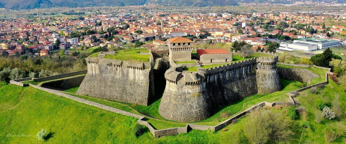 A historic castle surrounded by a fortified wall, set against a backdrop of a sprawling city and distant mountains.