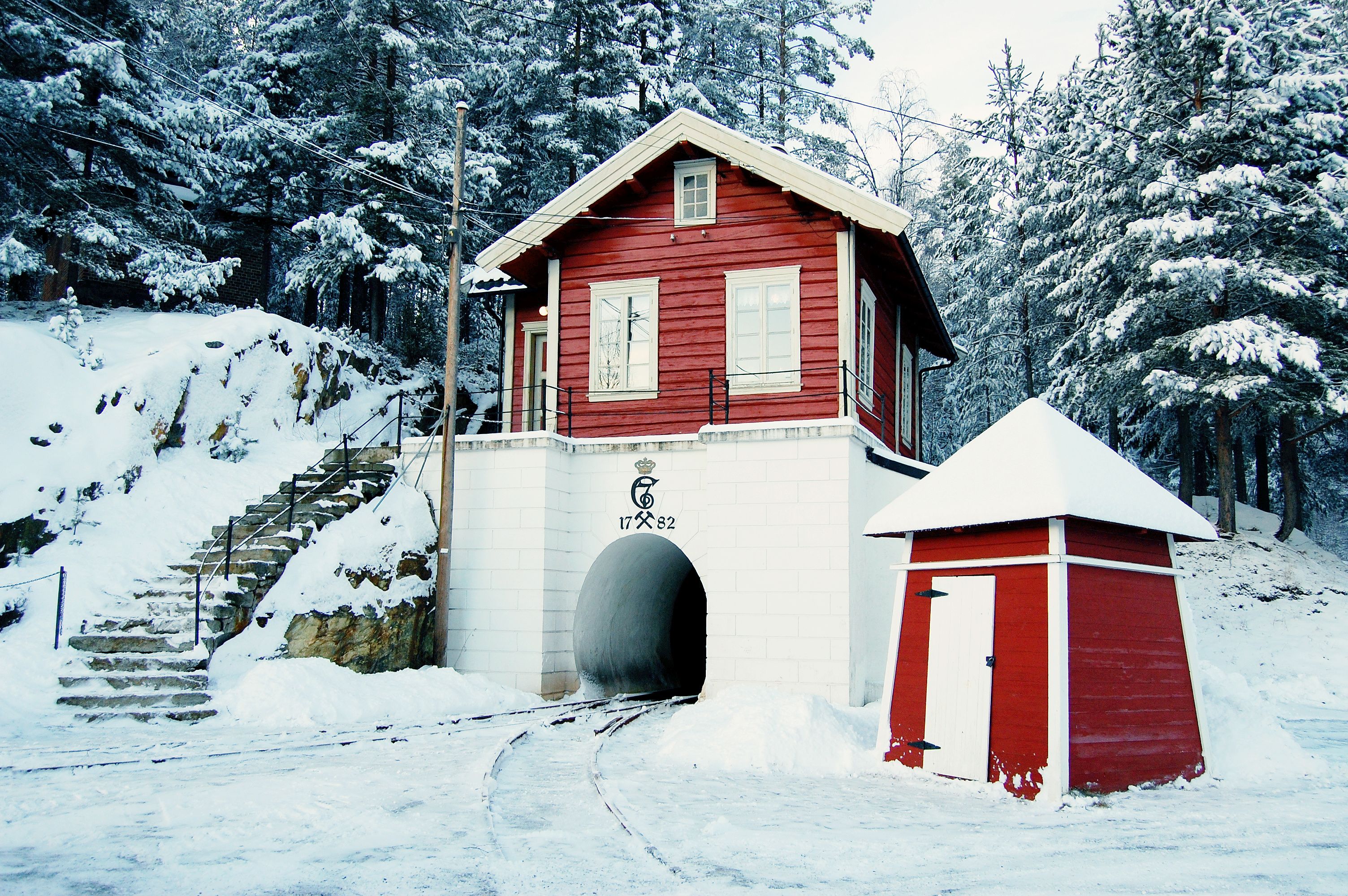 A red wooden cabin with a white door and a small shed, surrounded by snow-covered trees and a stone staircase leading up to the cabin.