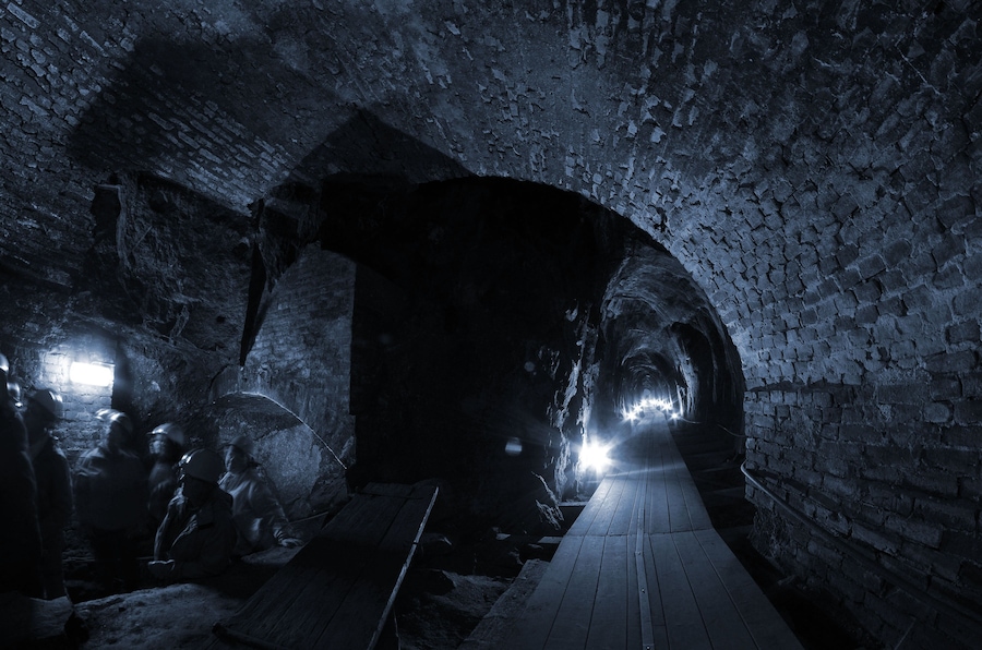 Un túnel subterráneo tenuemente iluminado, con paredes de ladrillo y tablones de madera en el suelo.