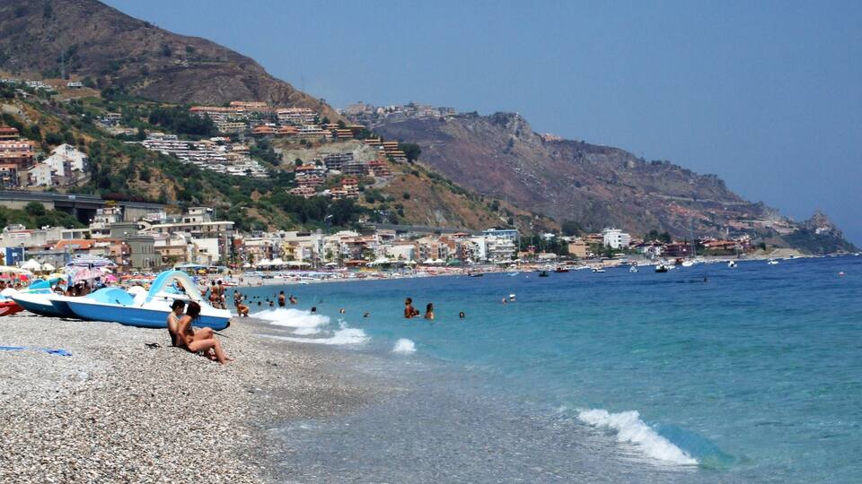A pebble beach with people and boats, a clear blue sea, and buildings on a hillside in the background.