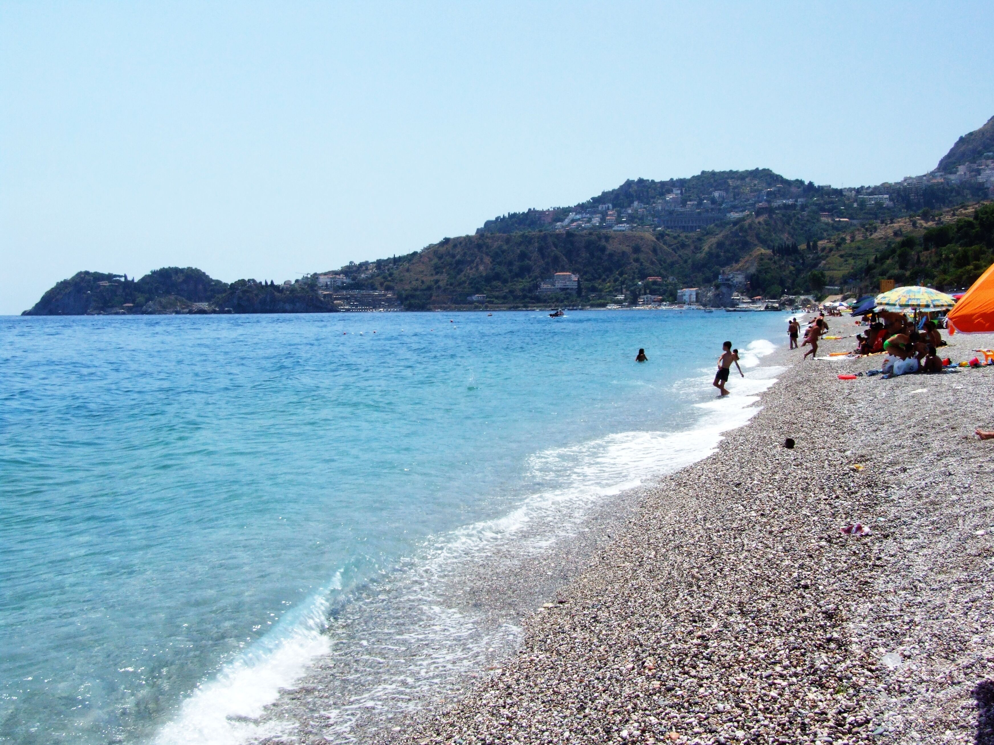 A pebble beach with clear blue water, a distant mountain, and a few people.
