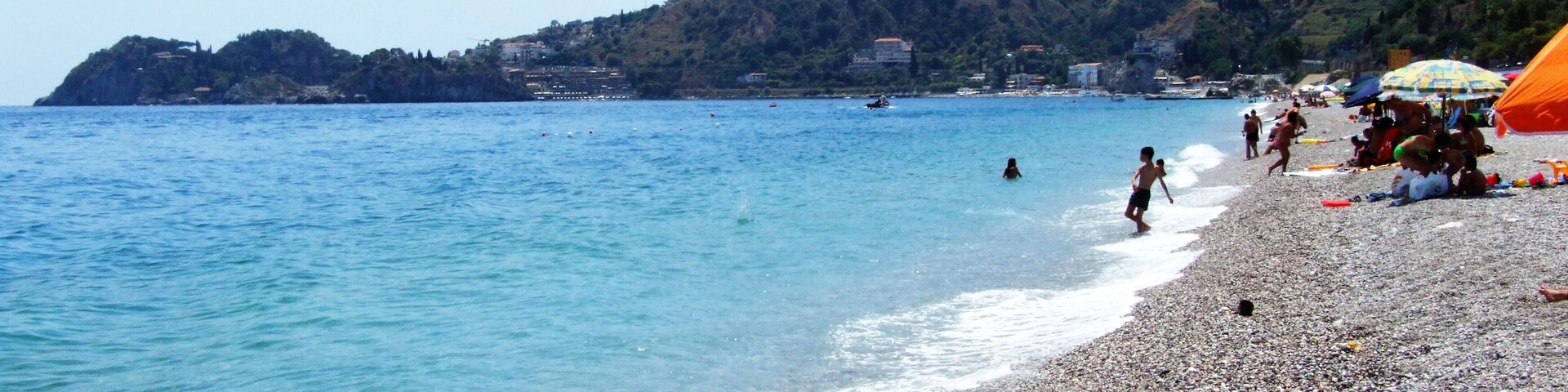 Uma praia de pedras com águas azul-claras, uma montanha ao longe e algumas pessoas.