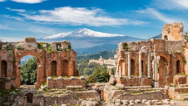 Ancient ruins with arches and columns, a snow-capped mountain in the background, and a town below.
