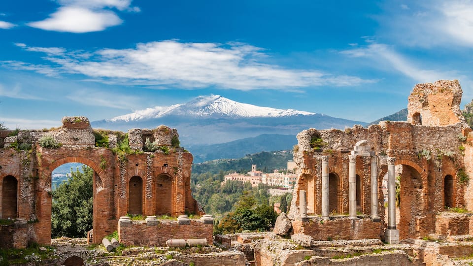 Ancient ruins with arches and columns, a snow-capped mountain in the background, and a town below.