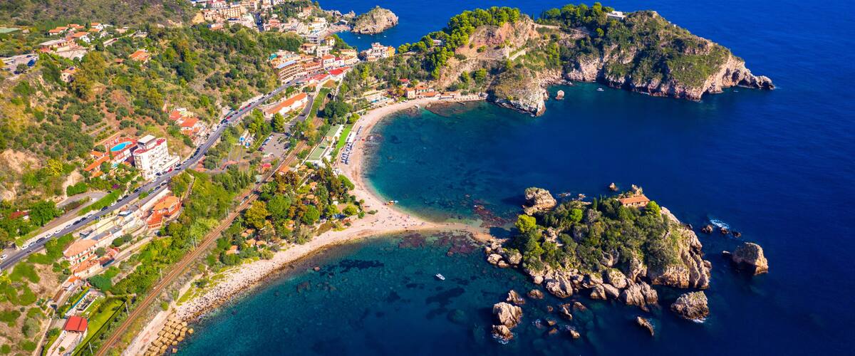 An aerial view of a coastal town with a beach, rocky outcrops, and a road leading to the town.