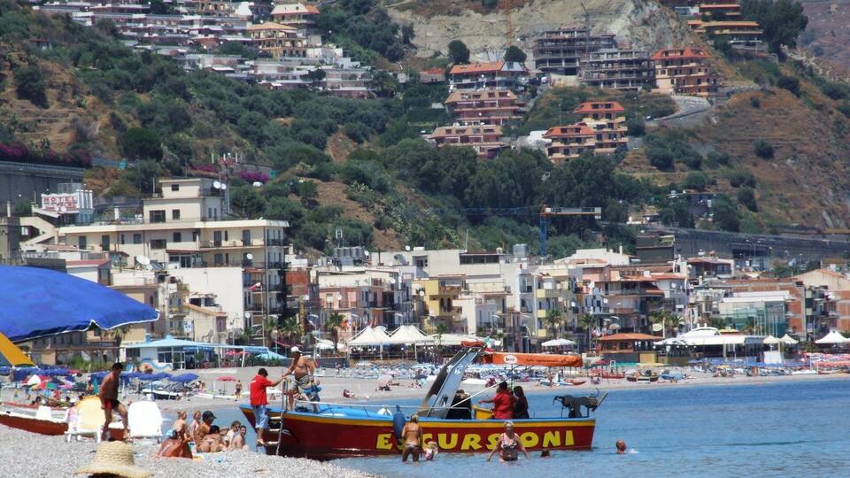 A coastal town with a beach, boats, and buildings on a hillside.
