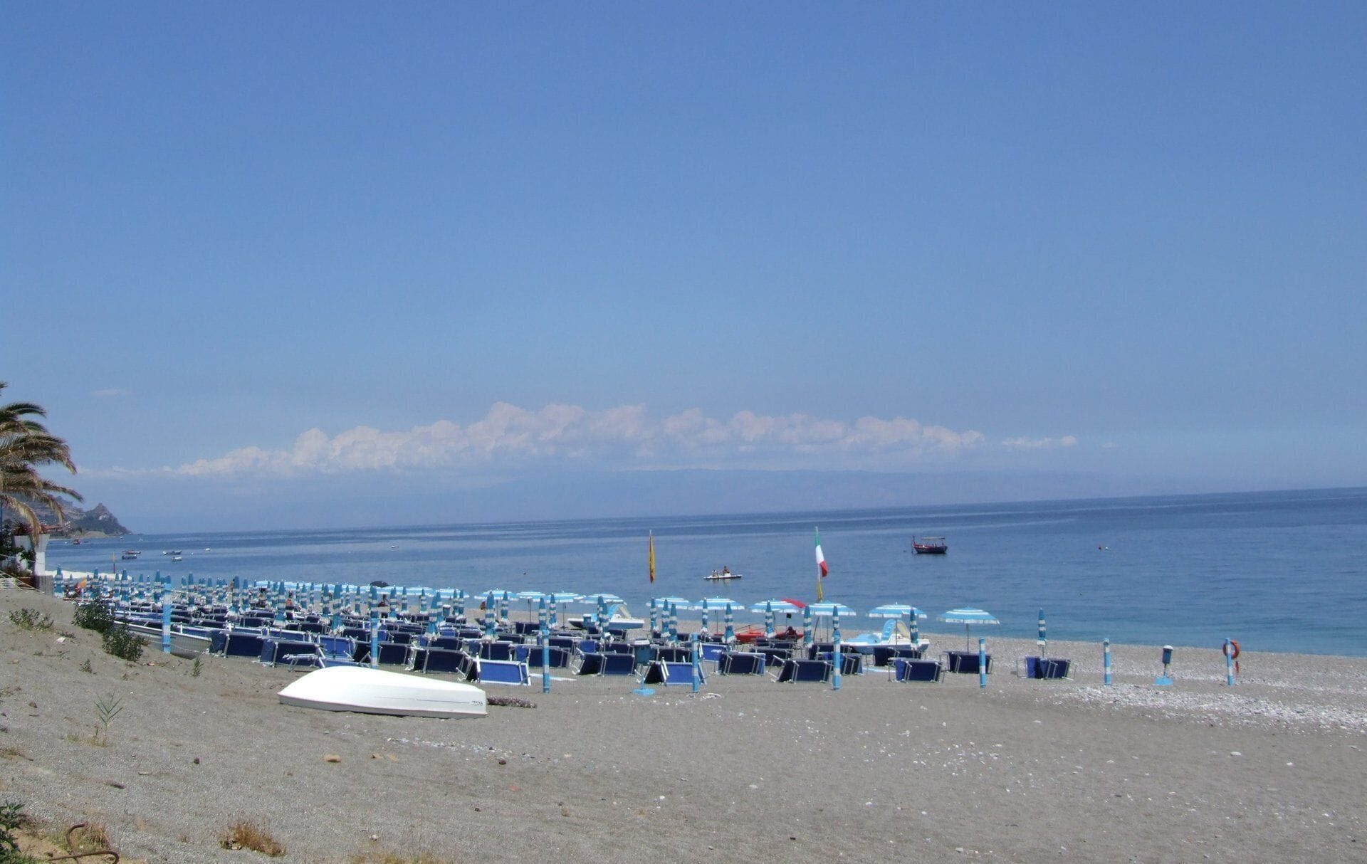 A beach with rows of blue beach chairs and umbrellas, a white boat, and a few boats on the water.
