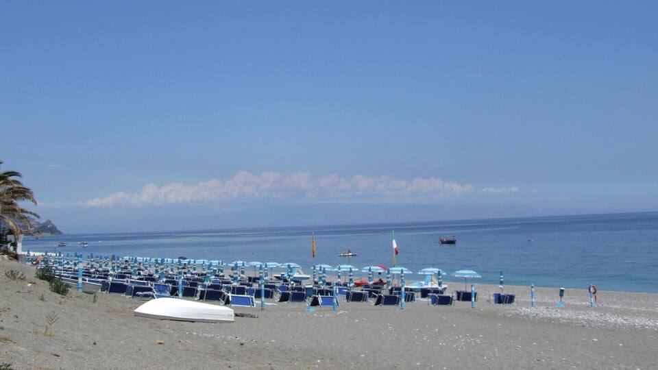 A beach with rows of blue beach chairs and umbrellas, a white boat, and a few boats on the water.