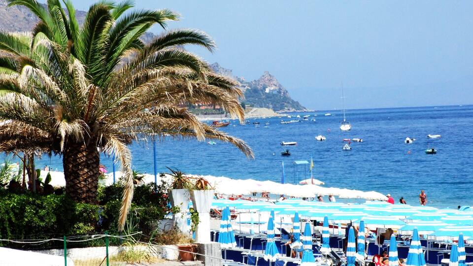 A beach with palm trees, blue umbrellas, and boats on the water.
