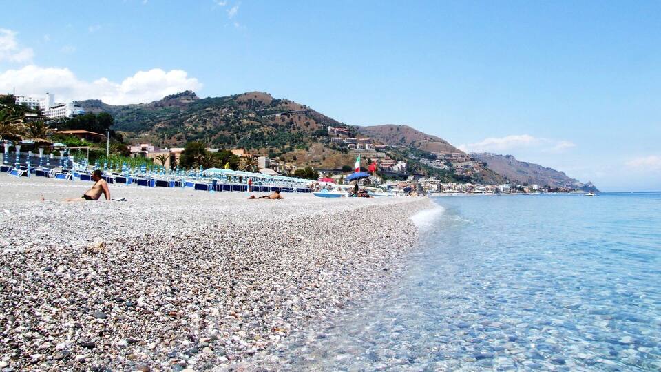 A pebble beach with clear water, a few people, and a town in the background.