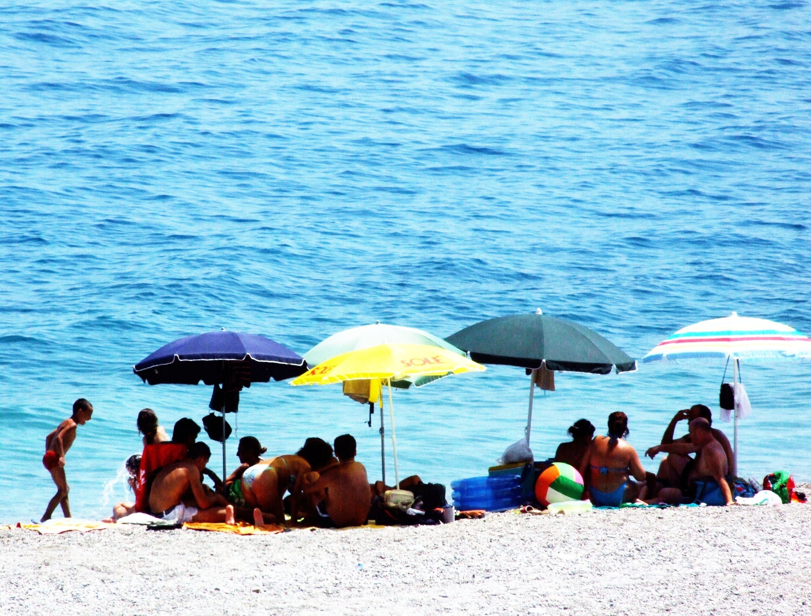 A beach with people relaxing under umbrellas and a volleyball on the sand.