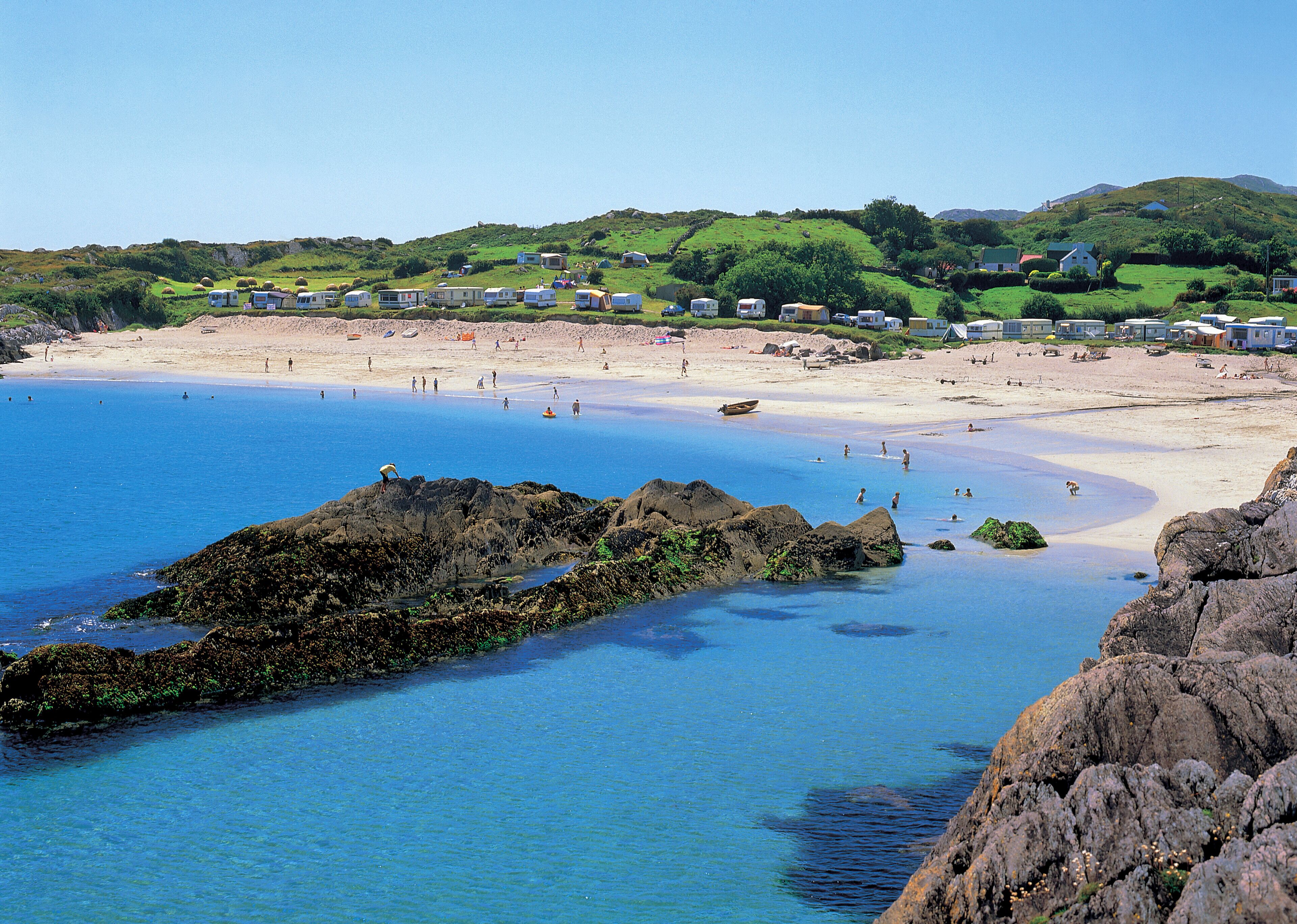 A coastal beach with clear blue waters, rocky outcrops, and a hilly landscape in the background.