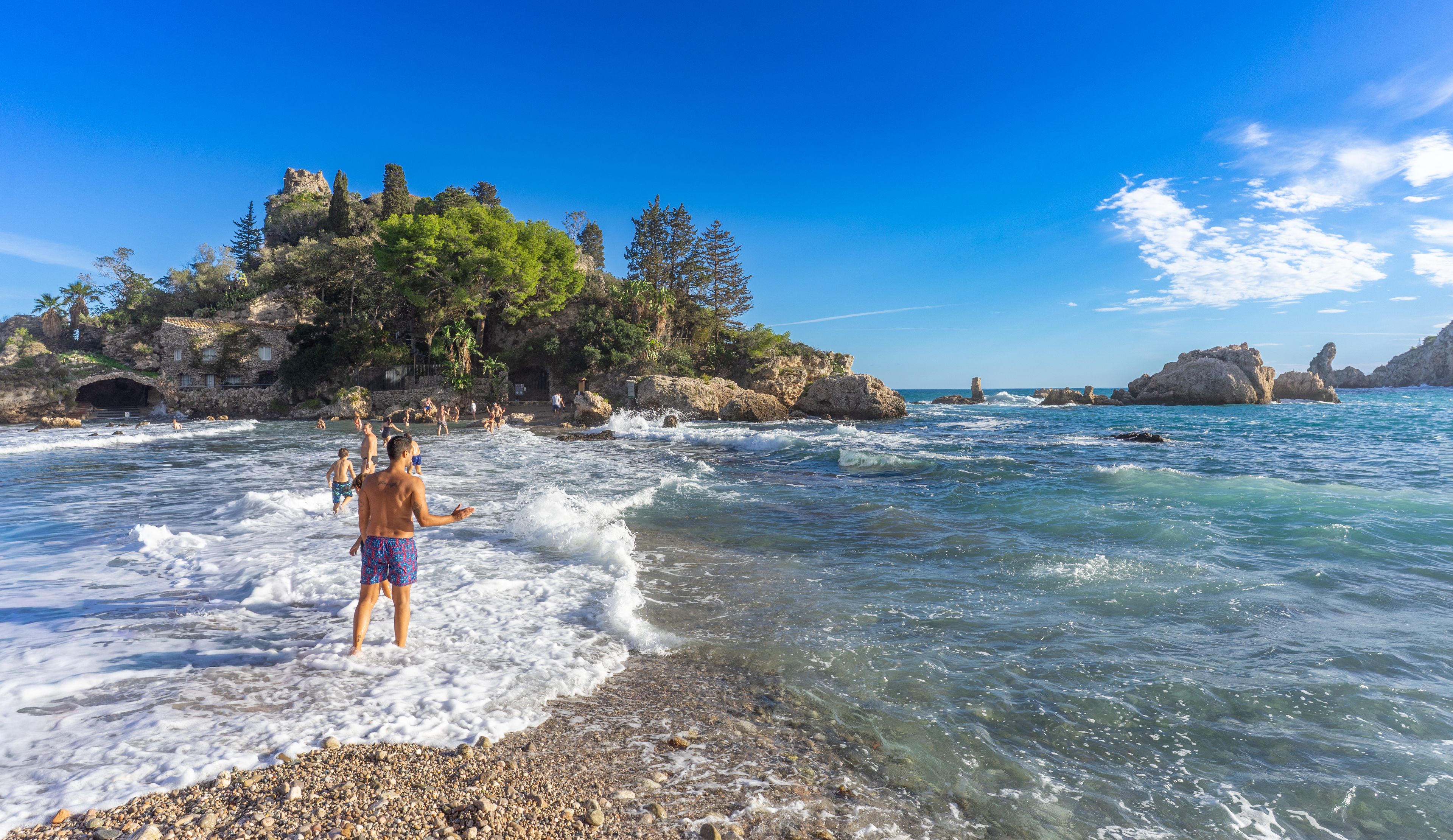 A beach with clear water, rocky shore, and a forested island in the background.