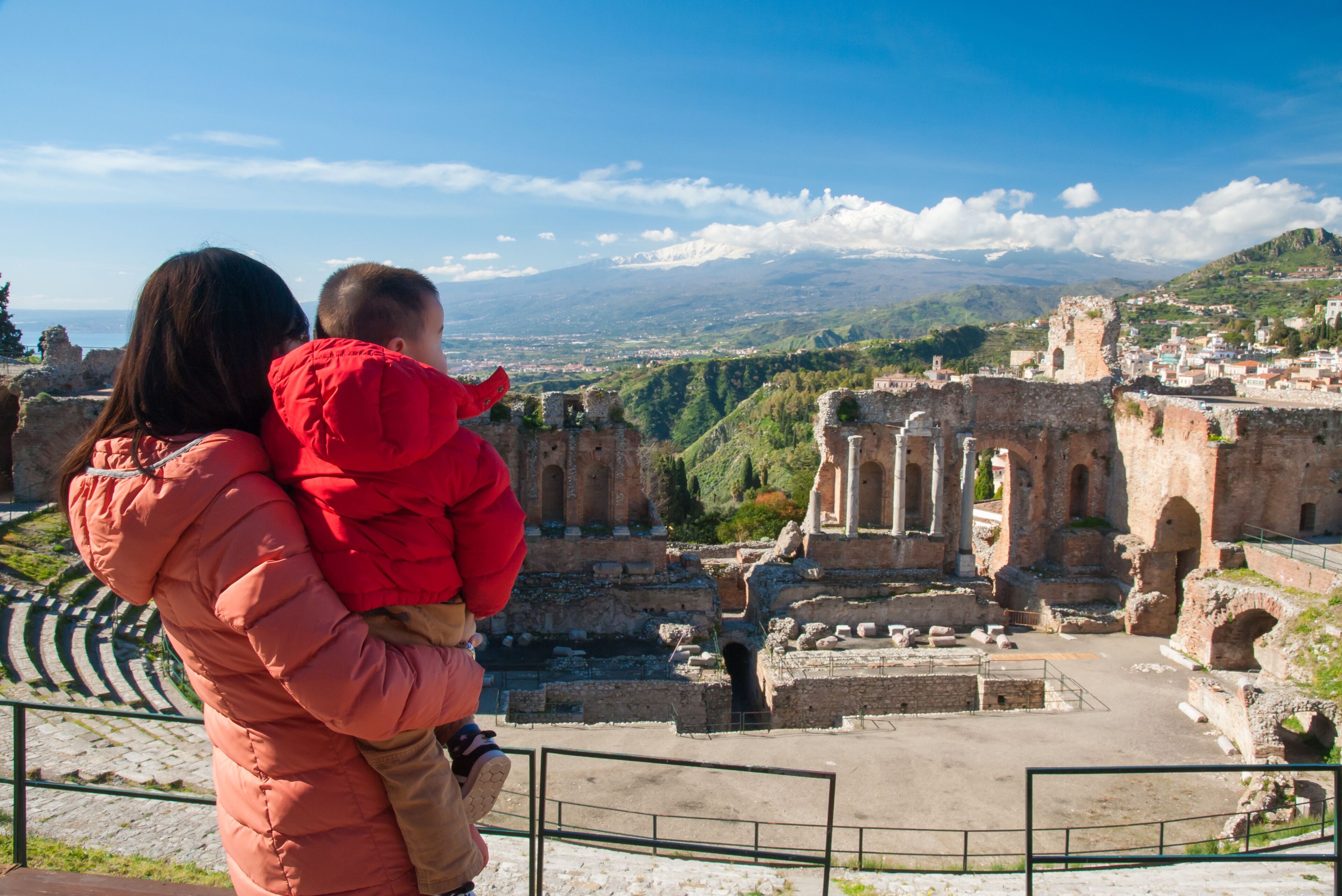 A person in a red jacket holding a child, overlooking ancient ruins with a mountainous landscape in the background.