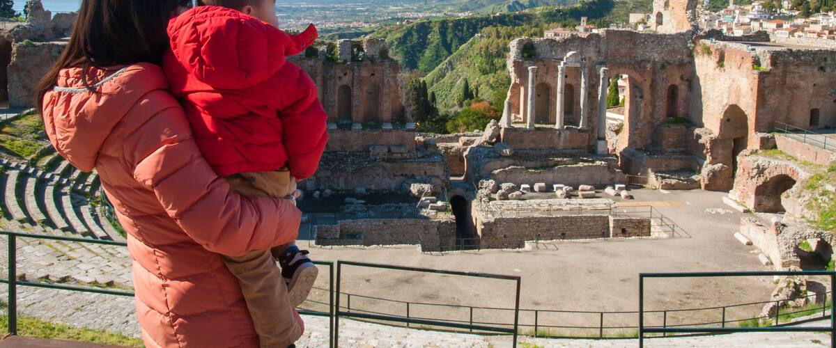A person in a red jacket holding a child, overlooking ancient ruins with a mountainous landscape in the background.