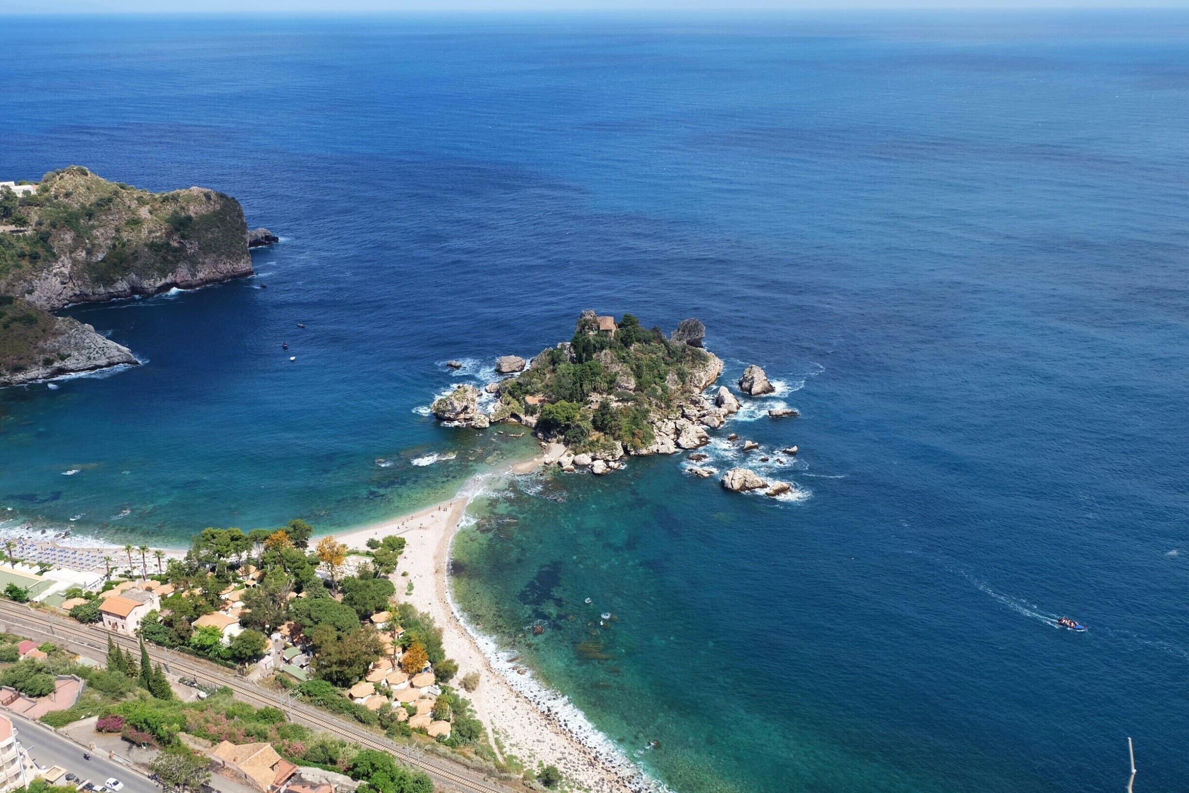 A coastal landscape with a rocky island, clear blue waters, and a beach with buildings and a road.