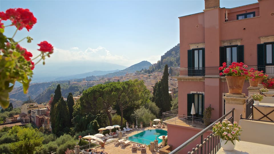 A poolside with lounge chairs and umbrellas, overlooking a hillside town with buildings and a mountain in the distance.