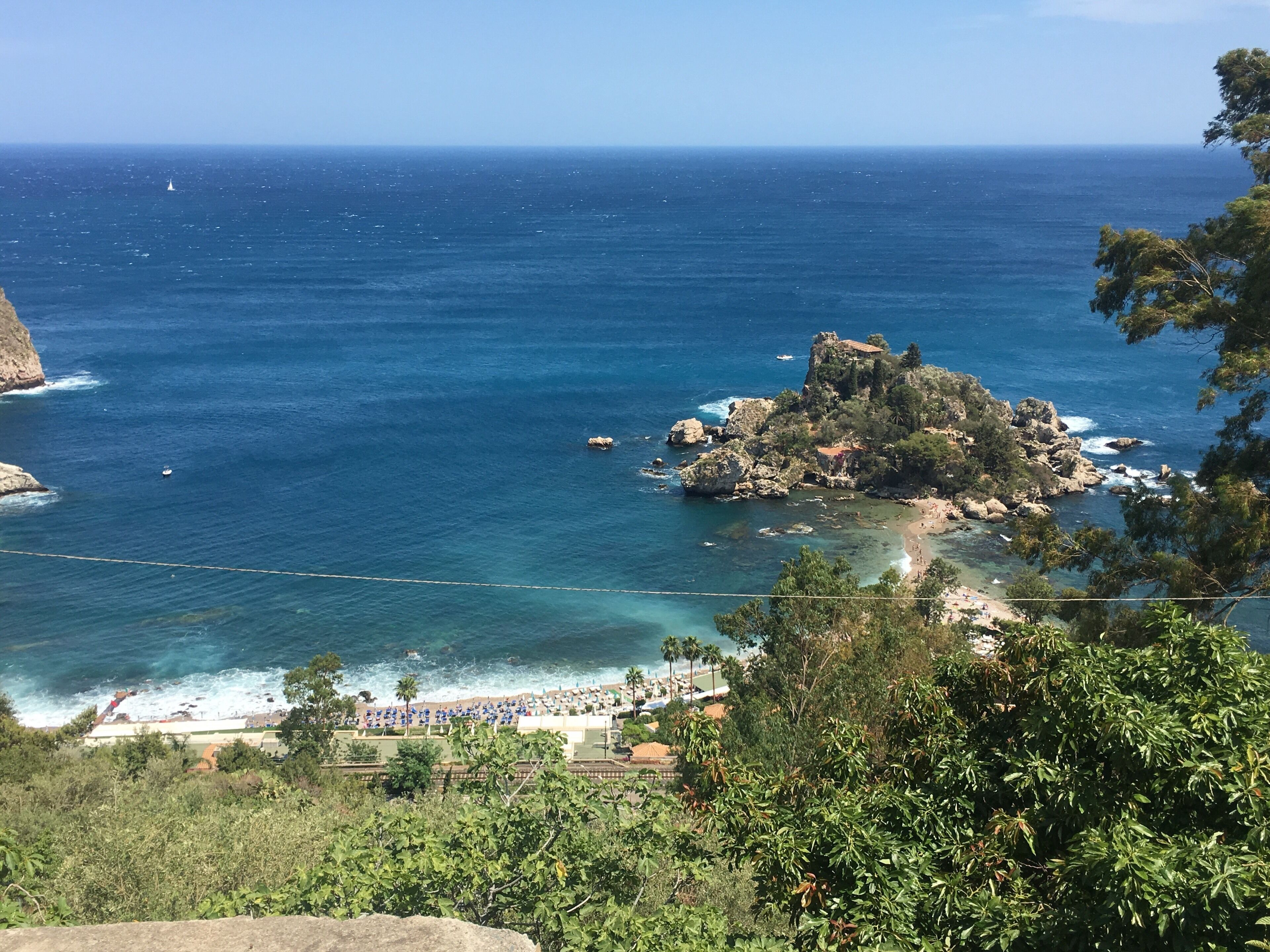 A coastal landscape with a rocky island, clear blue sea, and a beach area with umbrellas and people.