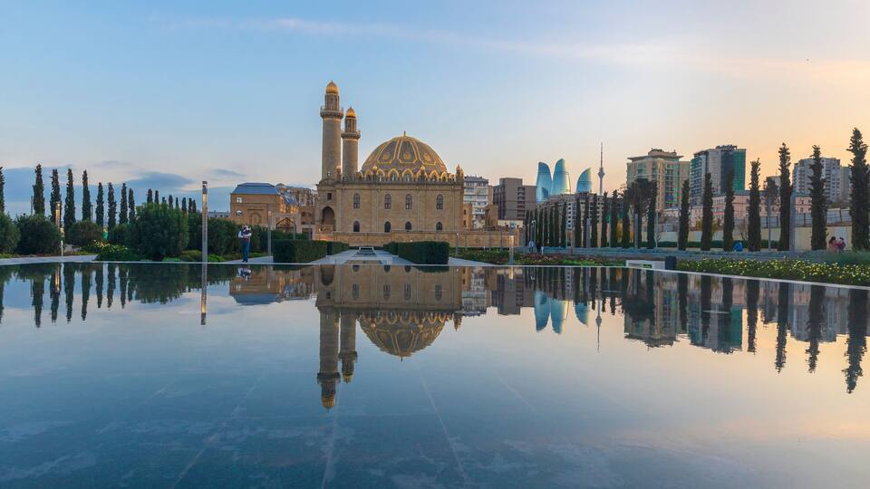 A mosque with a dome and minaret, reflecting in a still body of water, surrounded by modern buildings and trees.